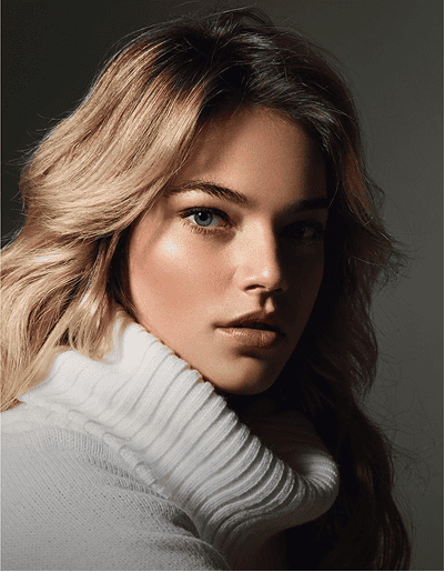 A woman with wavy blonde hair and blue eyes wears a white turtleneck sweater, looking directly at the camera against a neutral background.