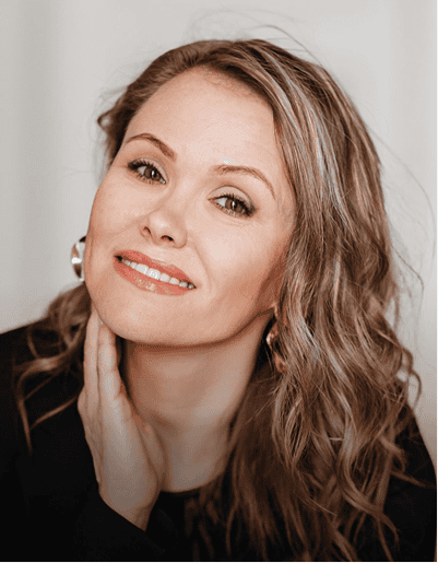 Woman with wavy, light brown hair and gold earrings, wearing a black top, tilts her head and smiles softly at the camera with her hand resting on her neck.