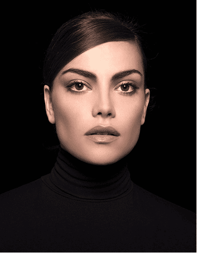 Woman with straight brown hair pulled back, wearing a black turtleneck, looking directly at the camera against a black background.