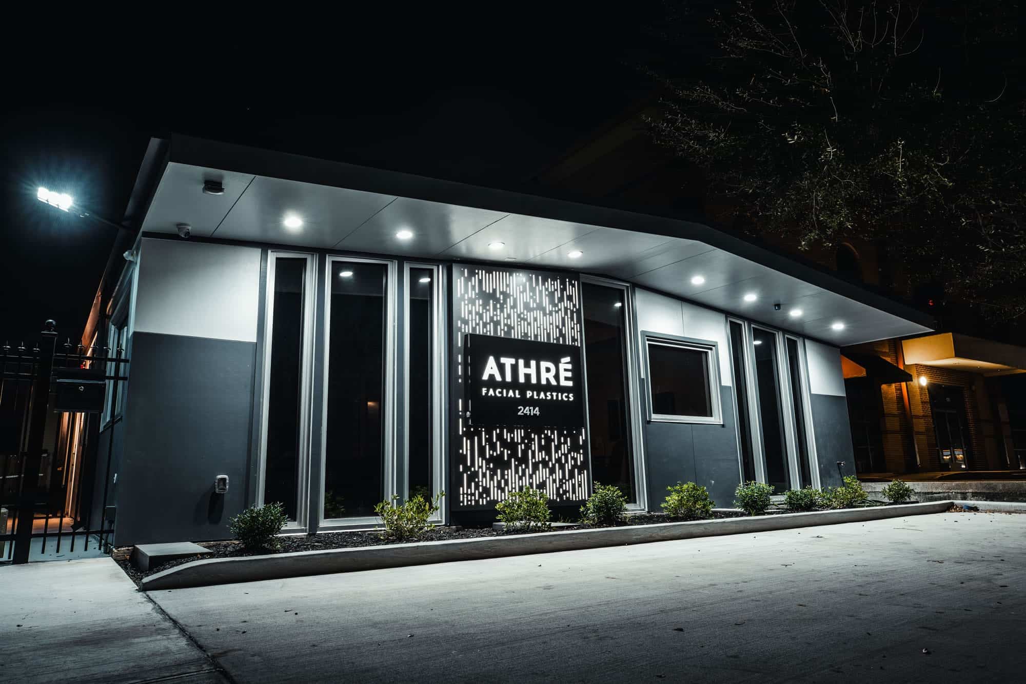 Modern commercial building with illuminated signage reading "ATHRÉ Facial Plastics 2414" at night, with exterior lights and landscaping visible.