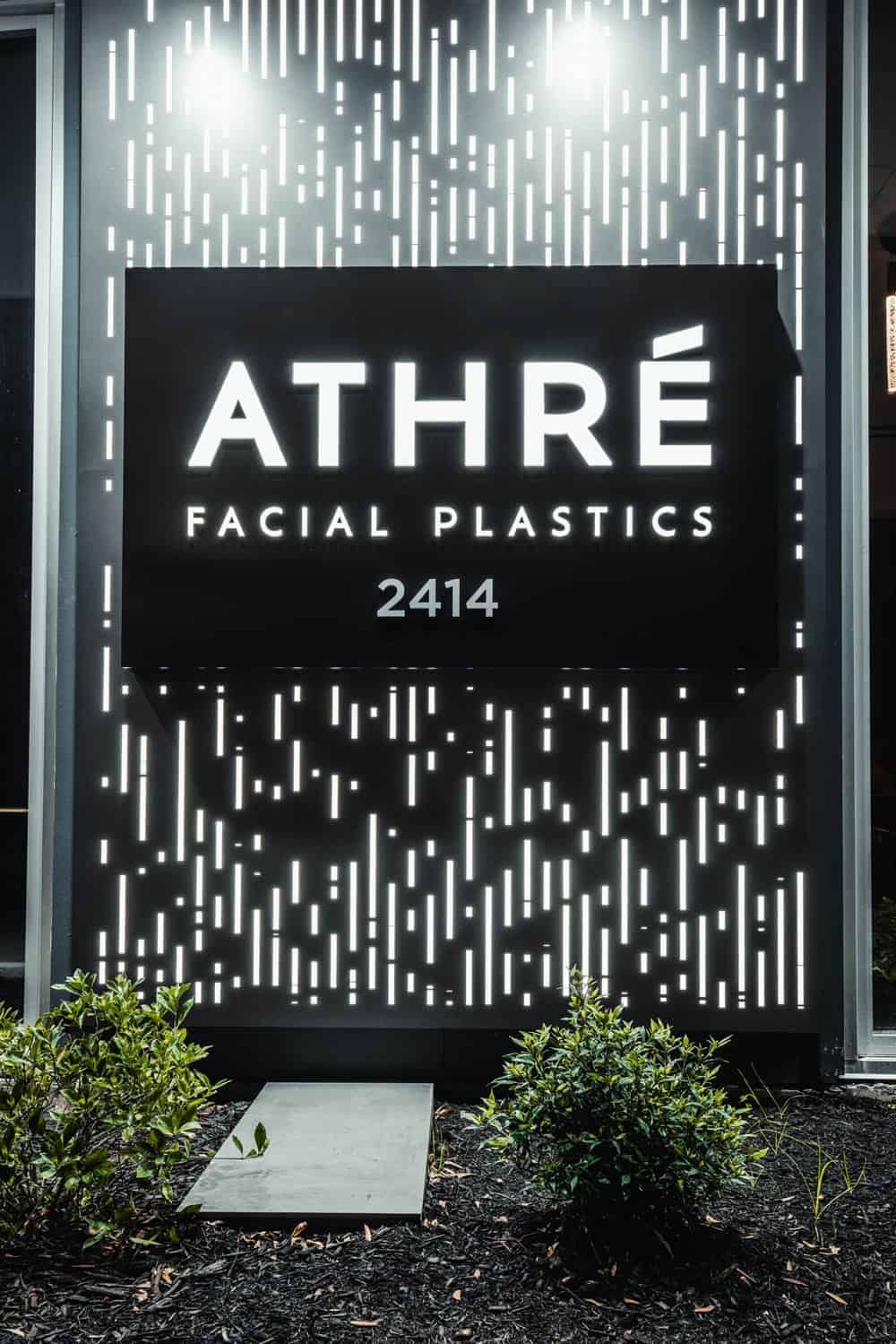 Black sign with white text reads "ATHRÉ FACIAL PLASTICS 2414" mounted on a wall with a decorative light pattern; small plants are in front of the sign.