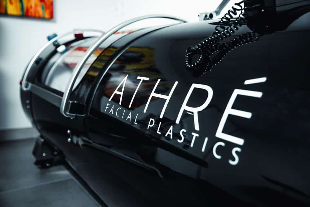 A black hyperbaric oxygen chamber with “ATHRÉ FACIAL PLASTICS” written on the side, in a modern medical office setting.