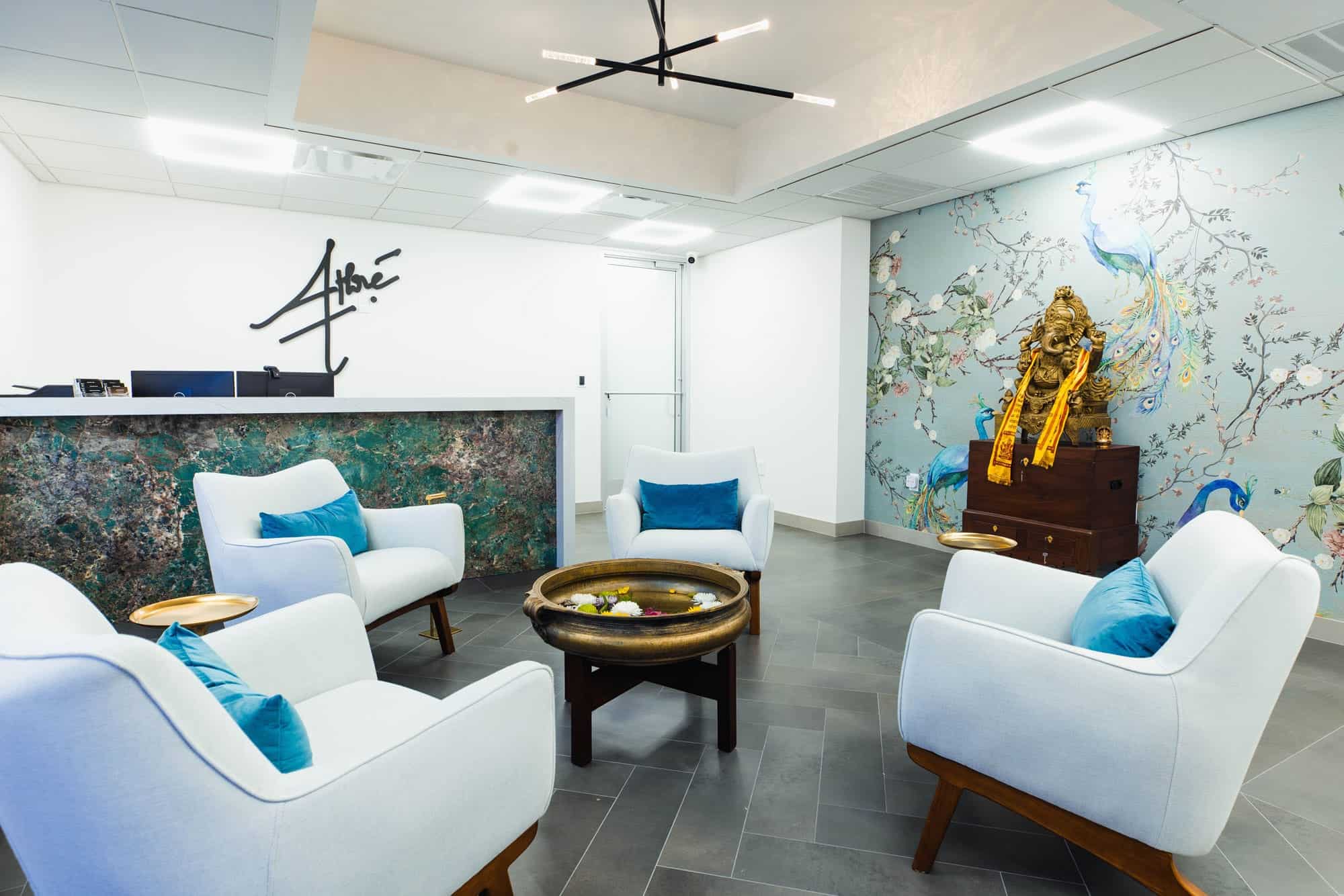 Modern waiting room with four white armchairs, a round table with a water bowl, a reception desk, and a wall mural featuring a peacock and flowers.