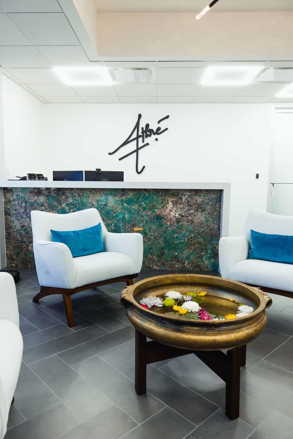 Modern office waiting area with white chairs, blue cushions, a round brass bowl with floating flowers, and a marble reception desk with the Athré logo on the white wall behind it.