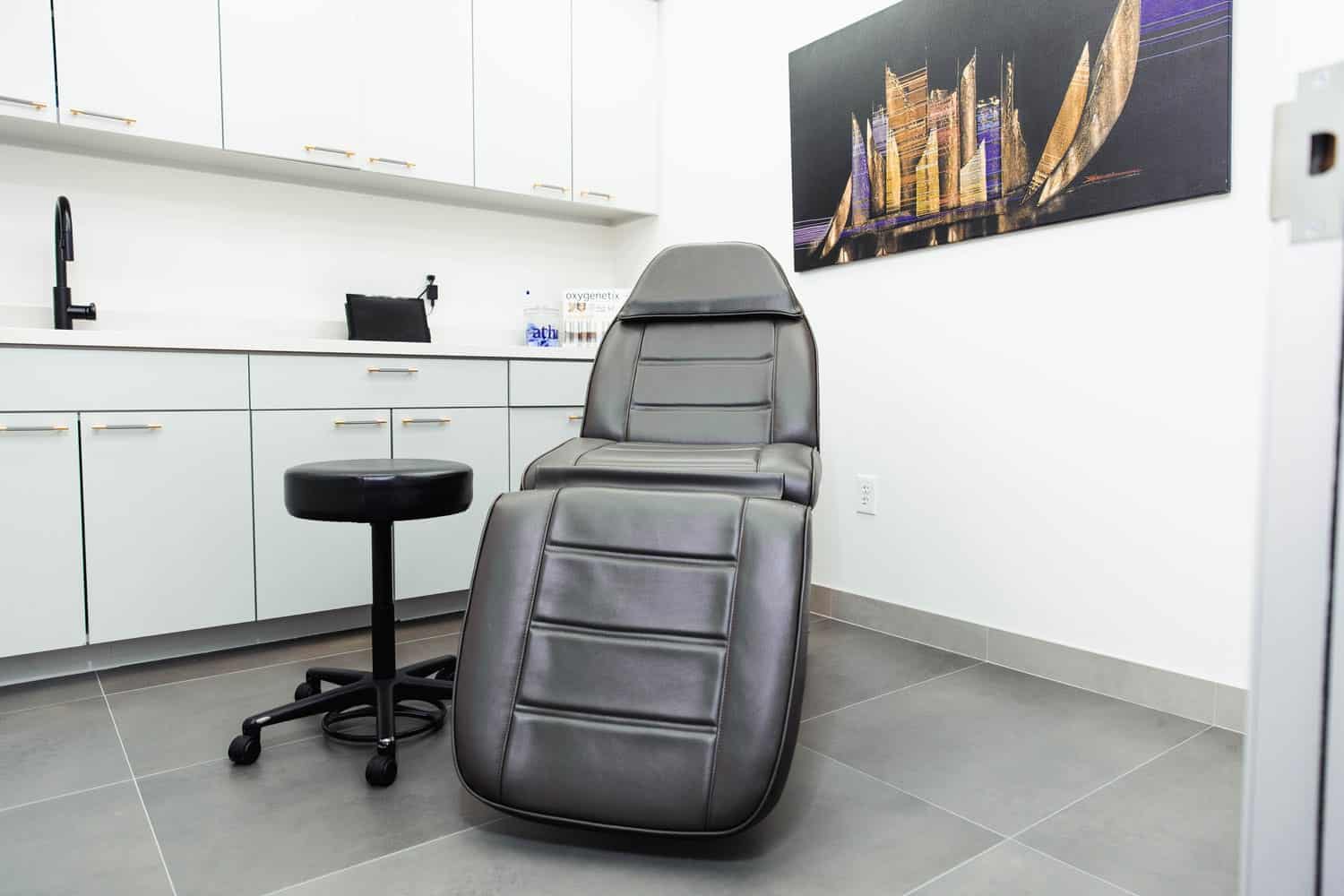 A black leather exam chair in a brightly lit facial plastic surgeon consultation room. An abstract painting hangs on the back wall.