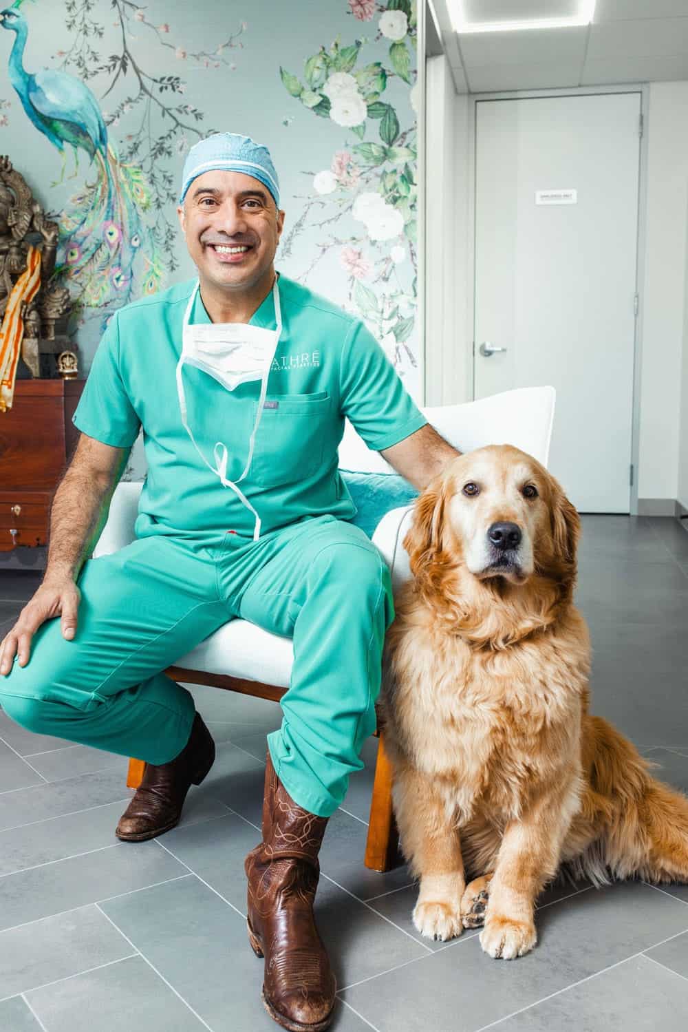 A man in teal medical scrubs and a surgical cap sits on a chair next to a golden retriever in a brightly lit room with floral wall art.