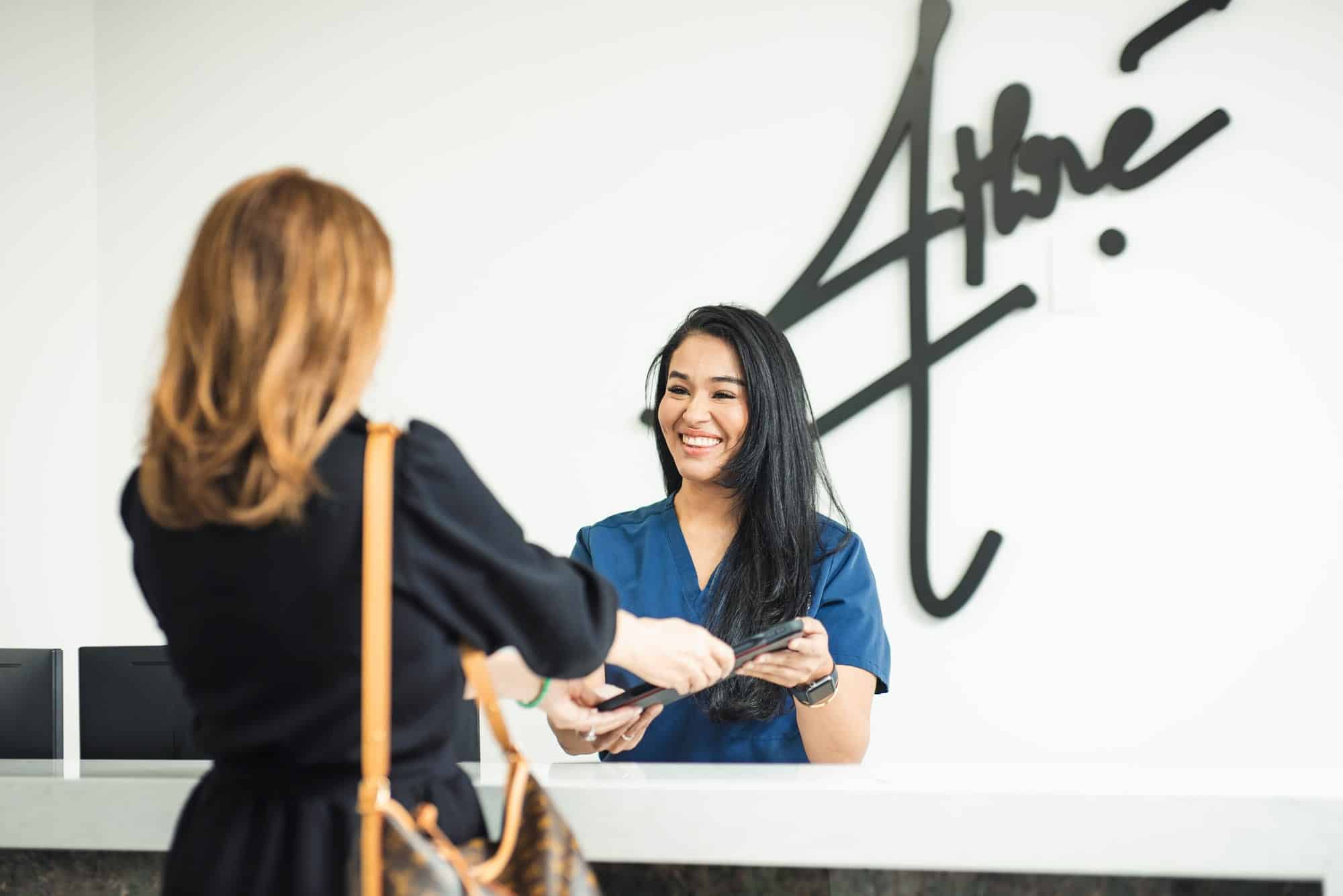 A woman in blue scrubs stands behind a reception desk, smiling and handing documents to another woman across the counter.