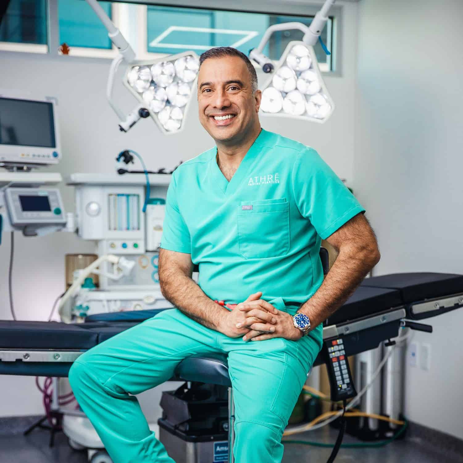 A man in teal medical scrubs sits on a stool in a brightly lit operating room, smiling at the camera with medical equipment visible in the background.