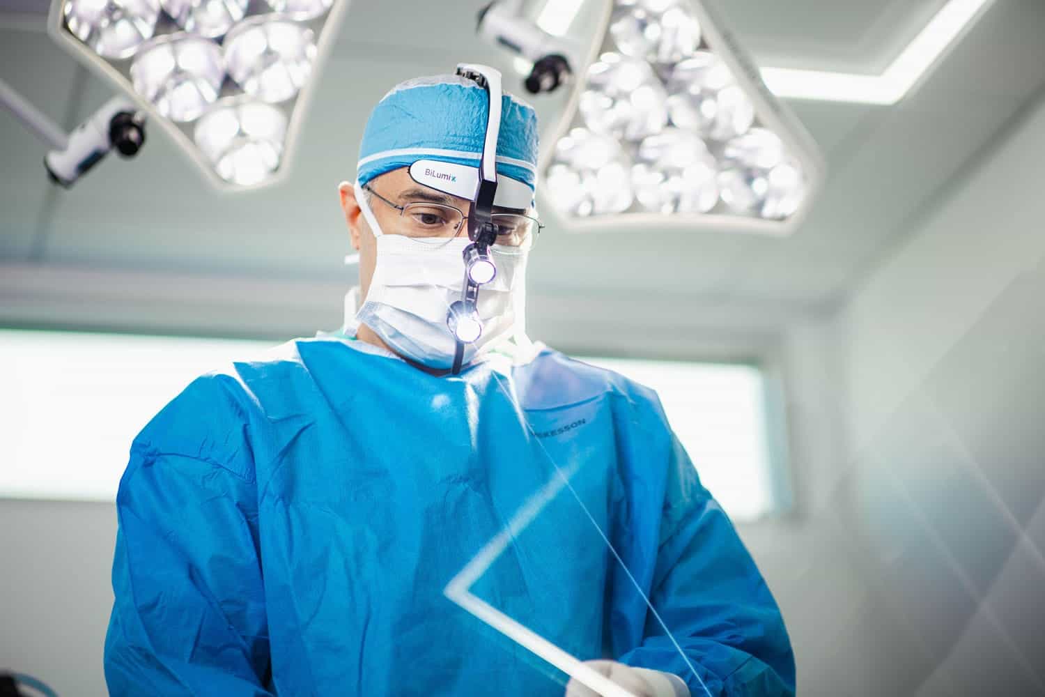 A surgeon in blue scrubs and mask, wearing magnifying glasses and a headlamp, operates under bright surgical lights in an operating room.