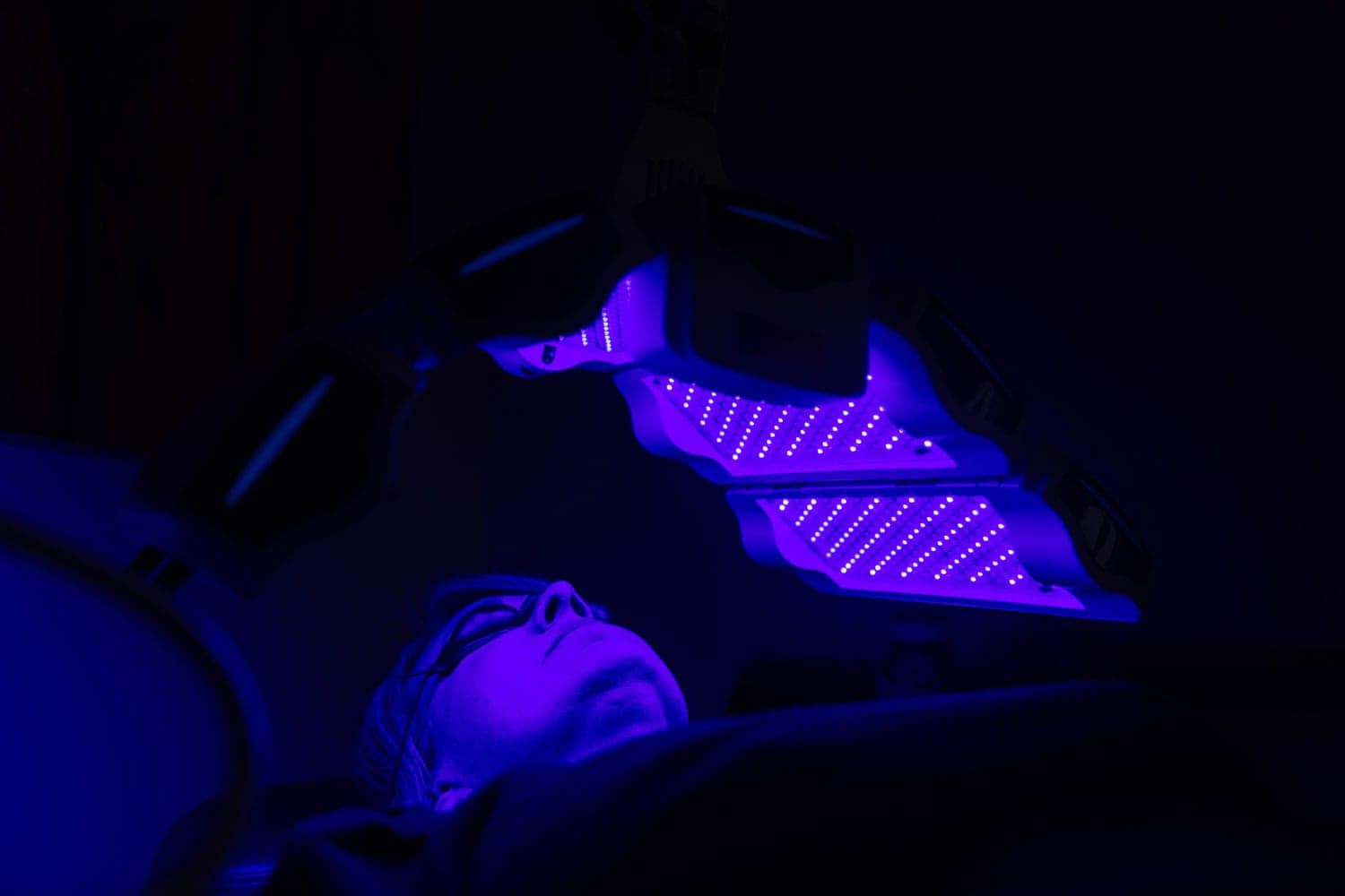 A person wearing protective goggles lies under a device emitting bright purple-blue LED lights in a dark room, receiving ElixirMD™ light therapy.