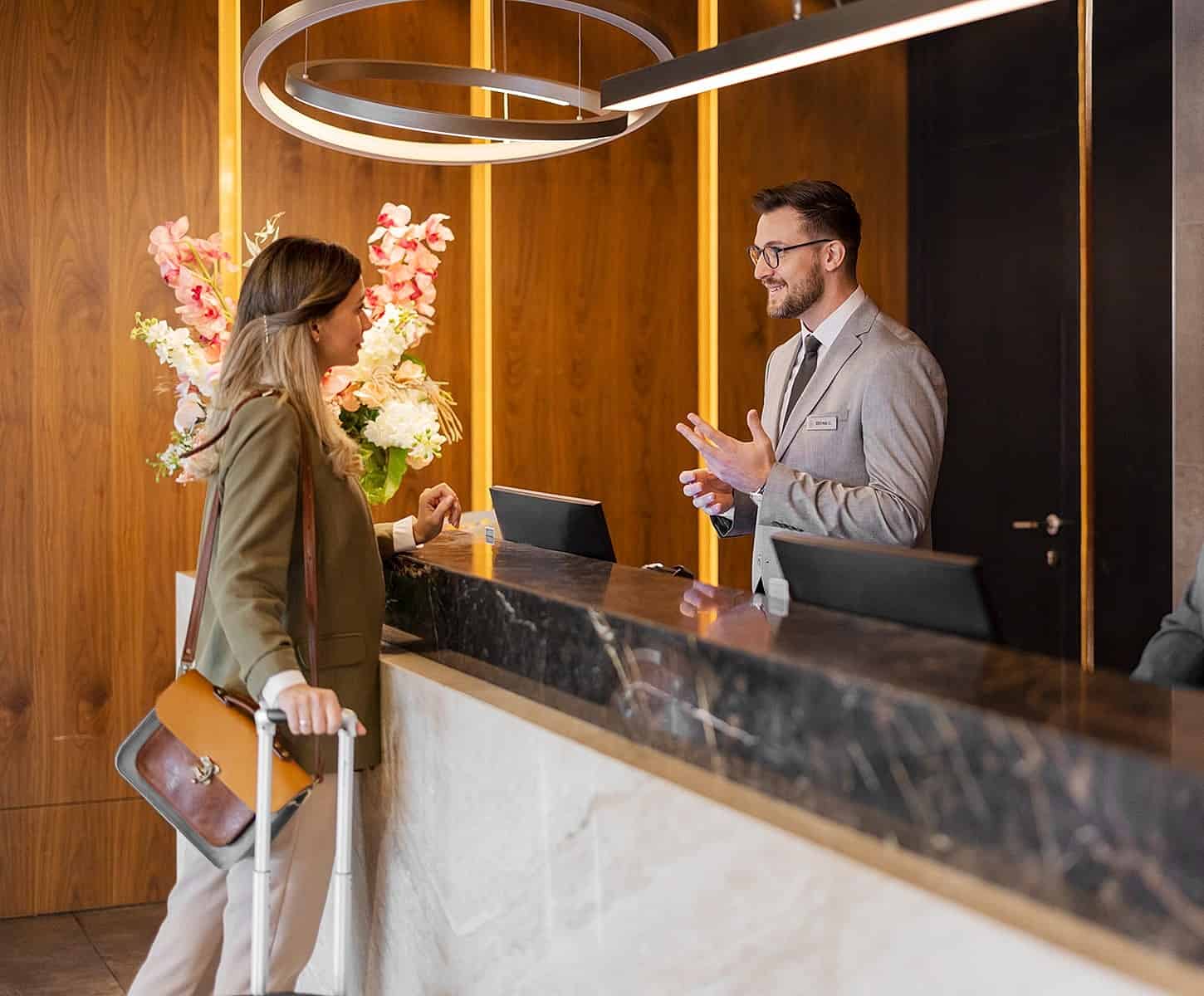 A woman with a suitcase speaks to a hotel receptionist at a modern front desk with flowers and wooden paneling in the background.