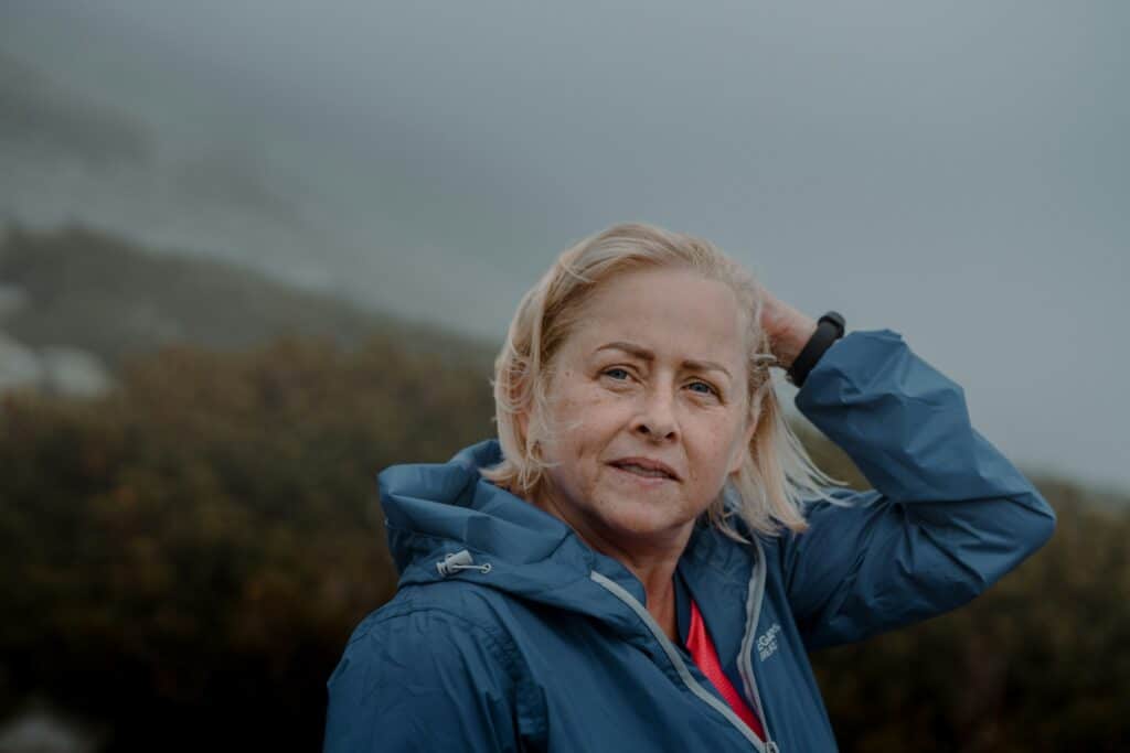 An older woman wearing a blue jacket stands outdoors on a foggy day.