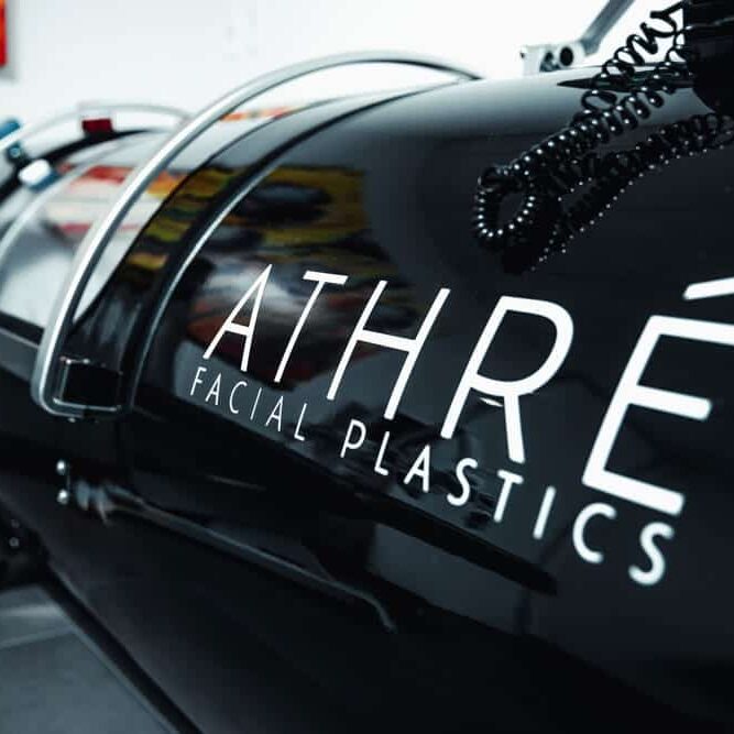 A black hyperbaric oxygen chamber with “ATHRÉ FACIAL PLASTICS” written on the side, in a modern medical office setting.