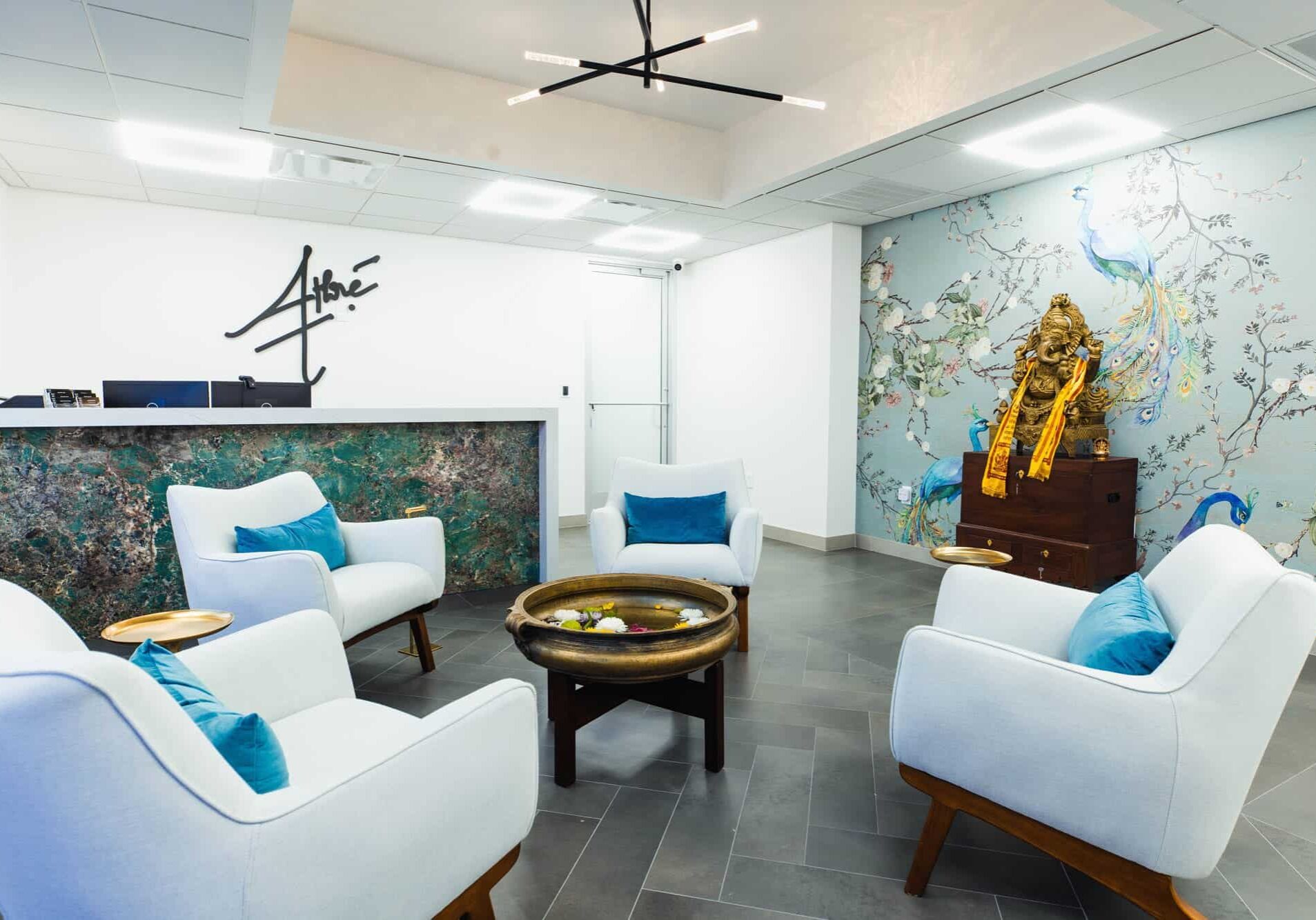 Modern waiting room with four white armchairs, a round table with a water bowl, a reception desk, and a wall mural featuring a peacock and flowers.
